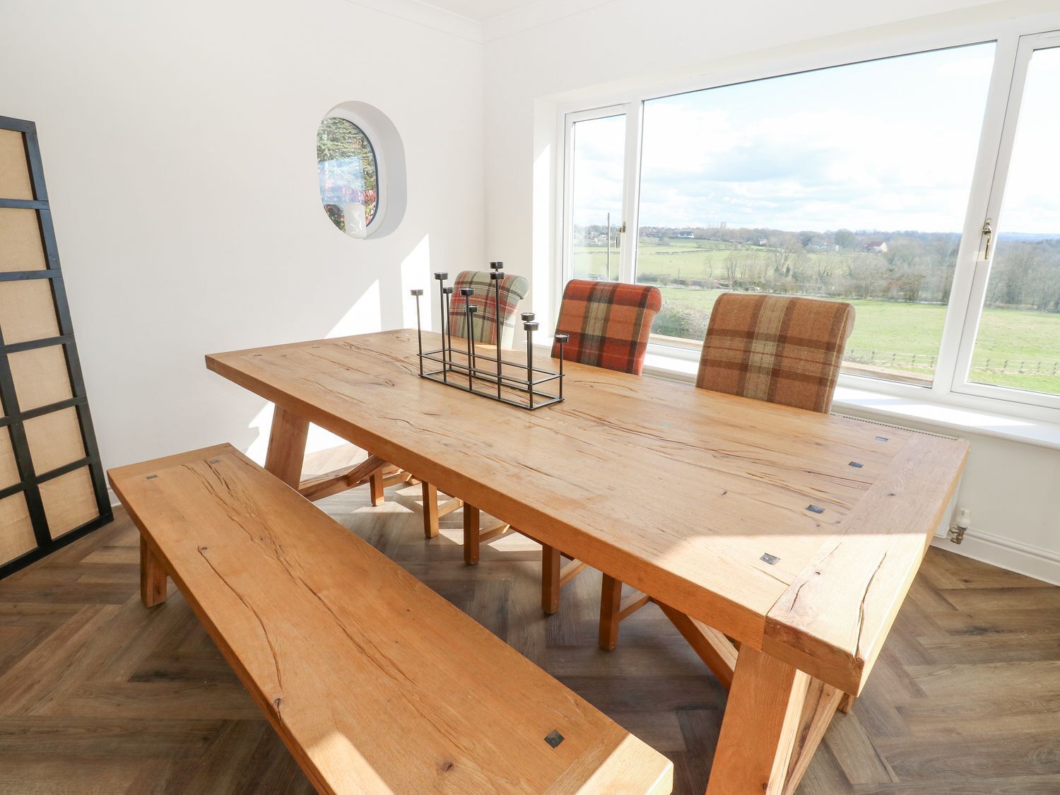 A wooden dining table with benches, plaid chairs, and a window with a view of a green landscape.