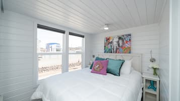 A bedroom with a large bed, a window with a view of the beach, and a colorful painting on the wall.