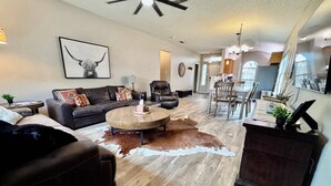 A spacious living room with a dark leather sofa, a round coffee table, and a cowhide rug. There is a dining area with wooden chairs and a chandelier. - Covered JM Ranch - 3BR/2BA - Family Friendly (Lubbock)