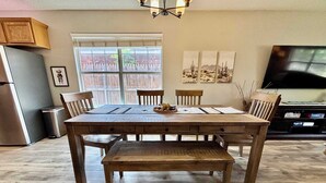 A dining area with a wooden table and chairs, a bench, a window with a view, a trash can, and a television. - Covered JM Ranch - 3BR/2BA - Family Friendly (Lubbock)