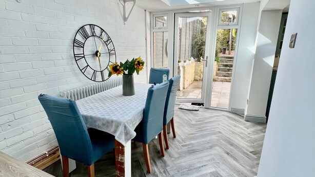 A dining area with a table, chairs, a clock, and a view through a skylight. - Housesleeps7-parking-garden-fireplace-beachnearby (Wareham)