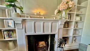 A fireplace with a white mantel, surrounded by shelves with books and decorative items. - Housesleeps7-parking-garden-fireplace-beachnearby (Wareham)