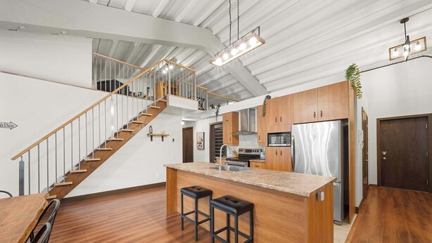 A modern kitchen with a central island, wooden flooring, a staircase, and a stainless steel refrigerator.