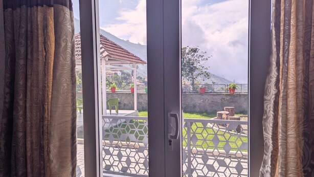 Vista attraverso una porta in vetro con tenda decorata, che si apre su un patio esterno con recinzione geometrica, piante in vaso e montagne sullo sfondo.