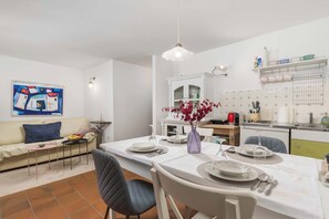 Un comedor con una mesa blanca dispuesta para cuatro personas, un florero con flores y una cocina al fondo.