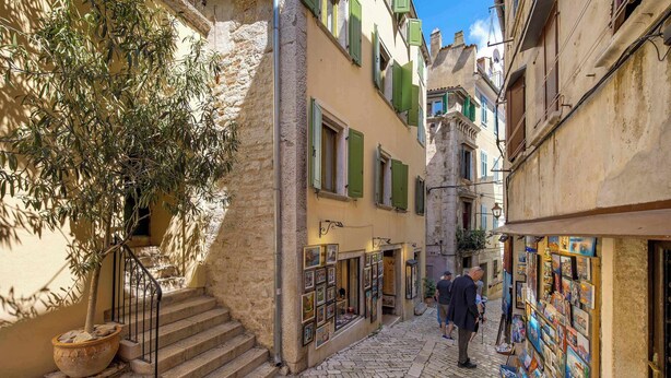 A narrow cobblestone street with buildings featuring traditional architecture, a staircase, and a shop with artwork displayed outside. - Apartment Dei Fiori 1 (Rovinj)