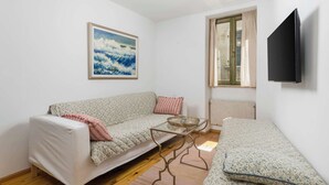 Ein Zimmer mit einer Couch, einem Glastisch, einem Bett, einem Gemälde, einem Fenster und einem Fernseher.