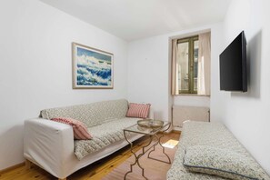 A room with a sofa, a glass coffee table, a bed, a painting, a window, and a TV.