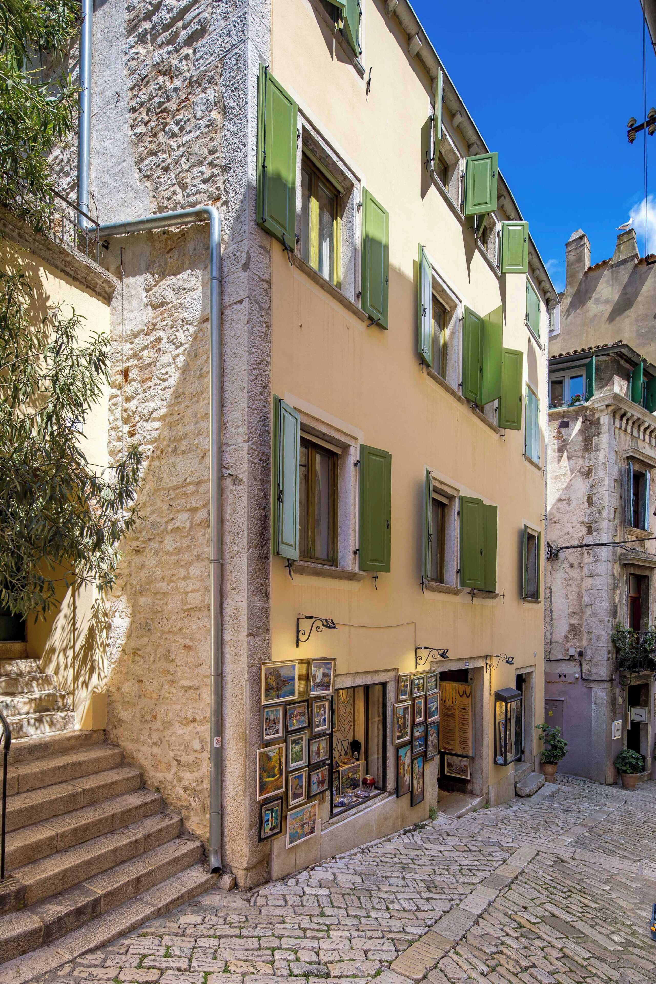 A narrow cobblestone street with a building featuring green shutters and a display of framed pictures.