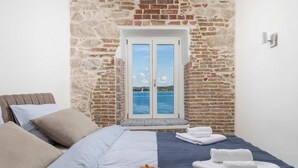 Une chambre avec un mur en pierre, une fenêtre donnant sur la mer, un lit avec des oreillers, une assiette de croissants et des serviettes pliées.