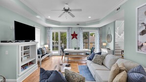 A spacious living room with a flat-screen TV, a ceiling fan, a dining area with a table and chairs, and a staircase leading to an upper level. - Rooftop Deck, Soaring Views & Steps to Tybee’s Best Beaches by Tybee Cottages (Tybee Island)