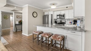 A modern kitchen with white cabinets, a stainless steel refrigerator, and a marble countertop. There are three wooden stools with leather seats. - Brighton 107 3 Br in Kingston Beach, Pool (Myrtle Beach)