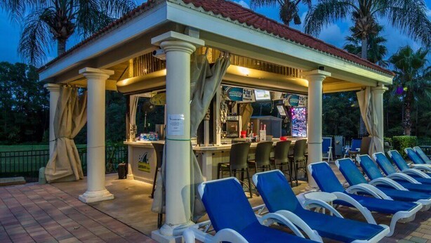 A covered outdoor seating area with lounge chairs, a bar, and palm trees in the background. - PROMO Villains Lair 8 BR Home, Private Pool - by Blue Sky Villas (Kissimmee)