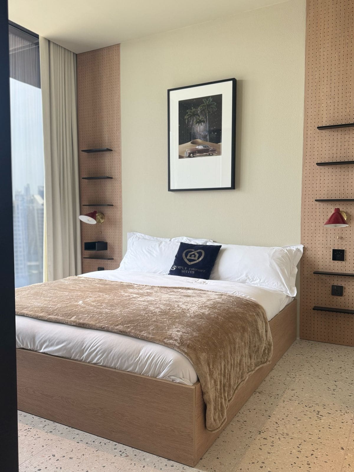 Habitació d'hotel amb llit, prestatge fixat a la paret, quadre amb marc i finestra amb cortines.