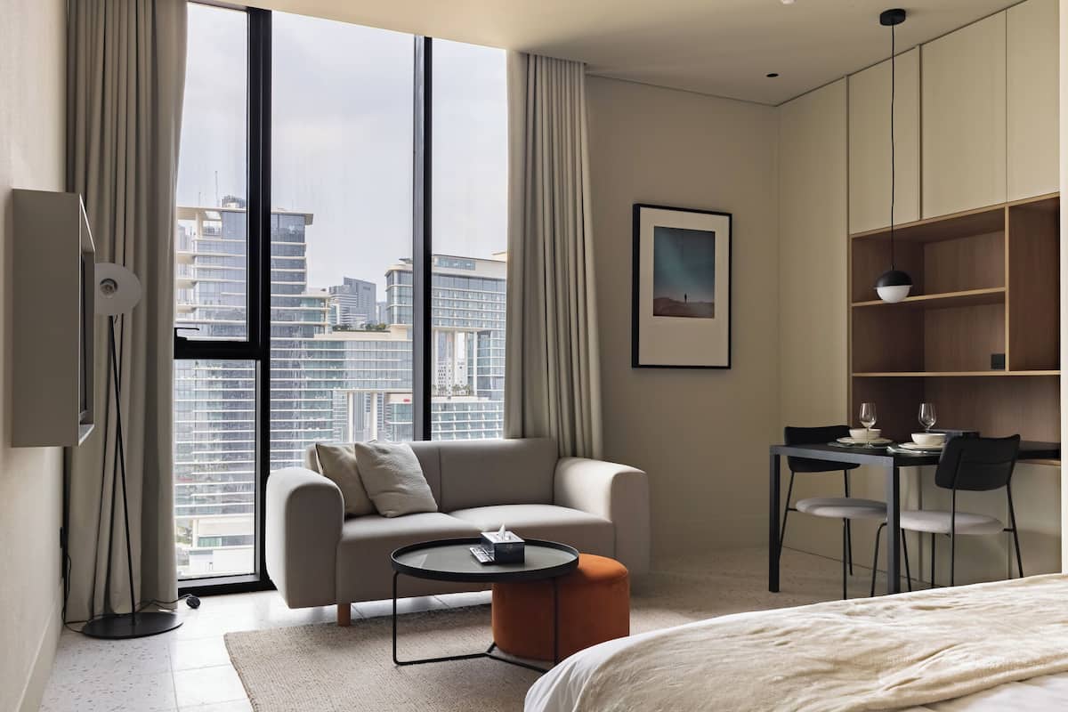 A modern hotel room with a large window, a sofa, a round coffee table, a bed, a desk with chairs, and a bookshelf.