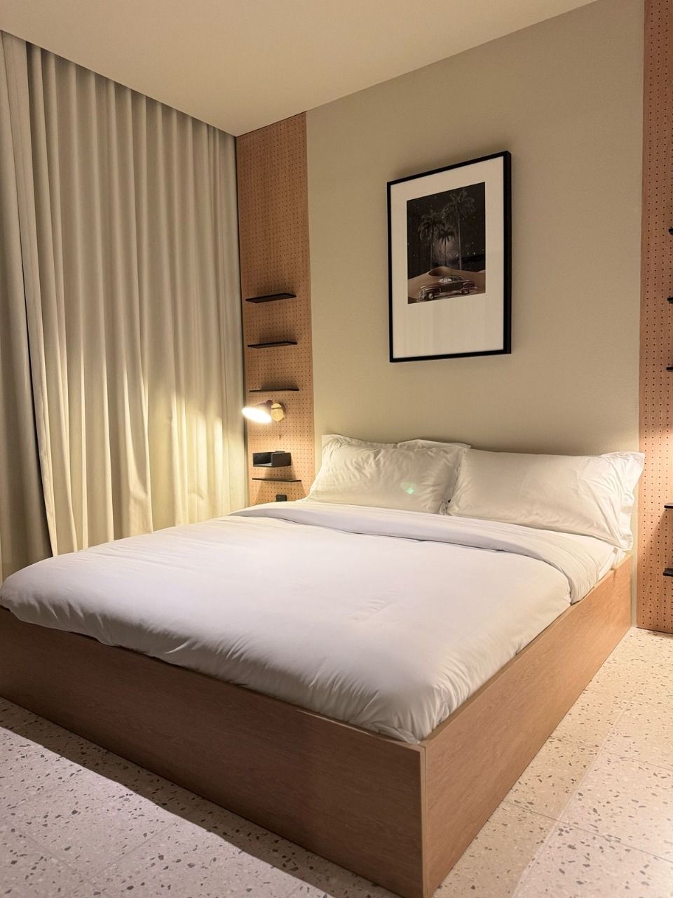 A modern hotel room with a large bed, a wall-mounted shelf, and a framed picture above the bed.