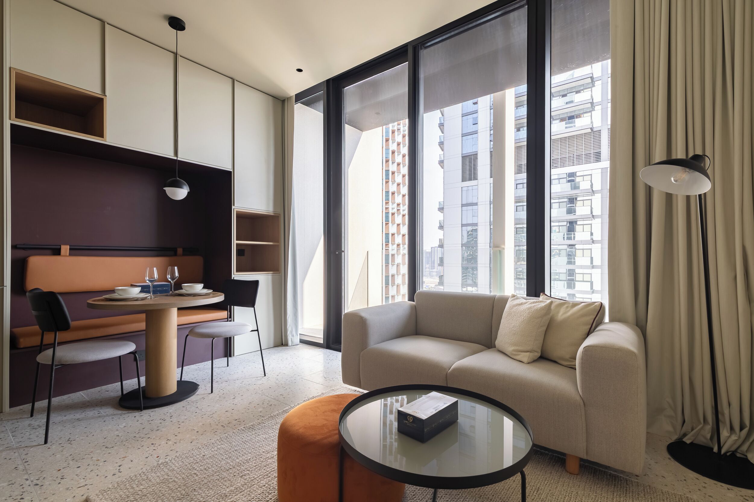 A modern hotel room with a sofa, a small round table, a dining area with a table and chairs, and large windows offering a city view.