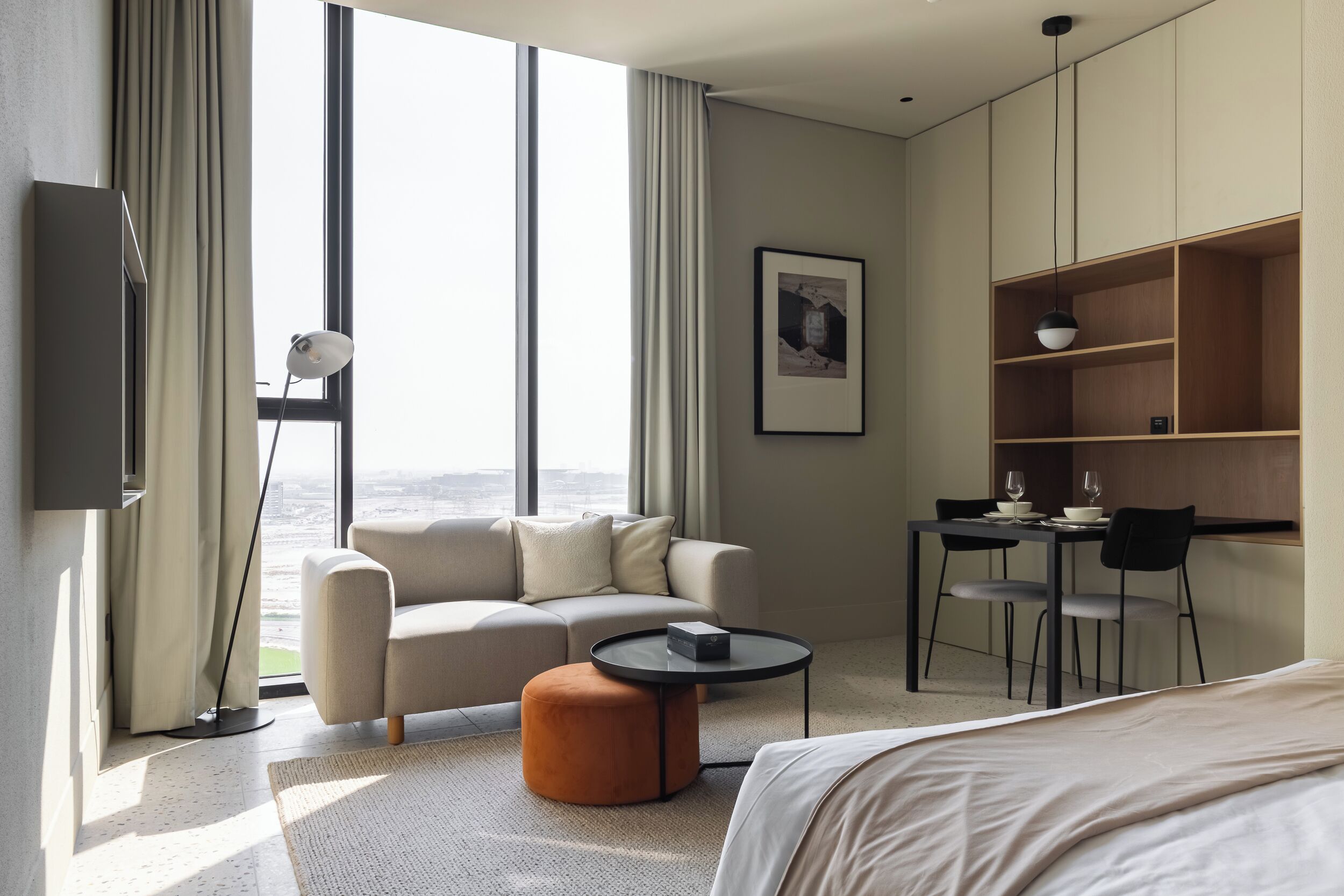 A modern hotel room with a large window, a sofa, a round coffee table, a small ottoman, a bed, a desk with chairs, and a bookshelf.