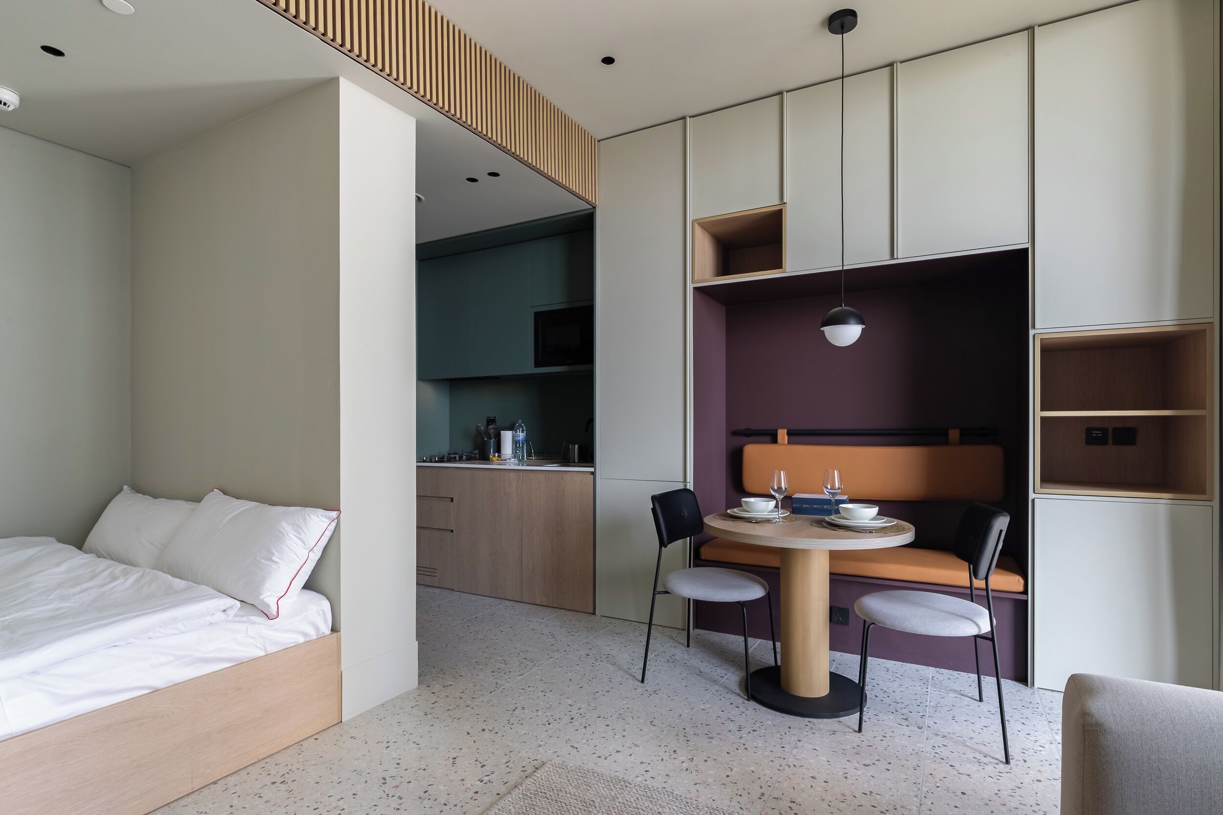 Ein modernes Hotelzimmer mit einem Bett, einer Essecke mit Tisch und Stühlen, einer Küchenzeile und integrierten Regalen.