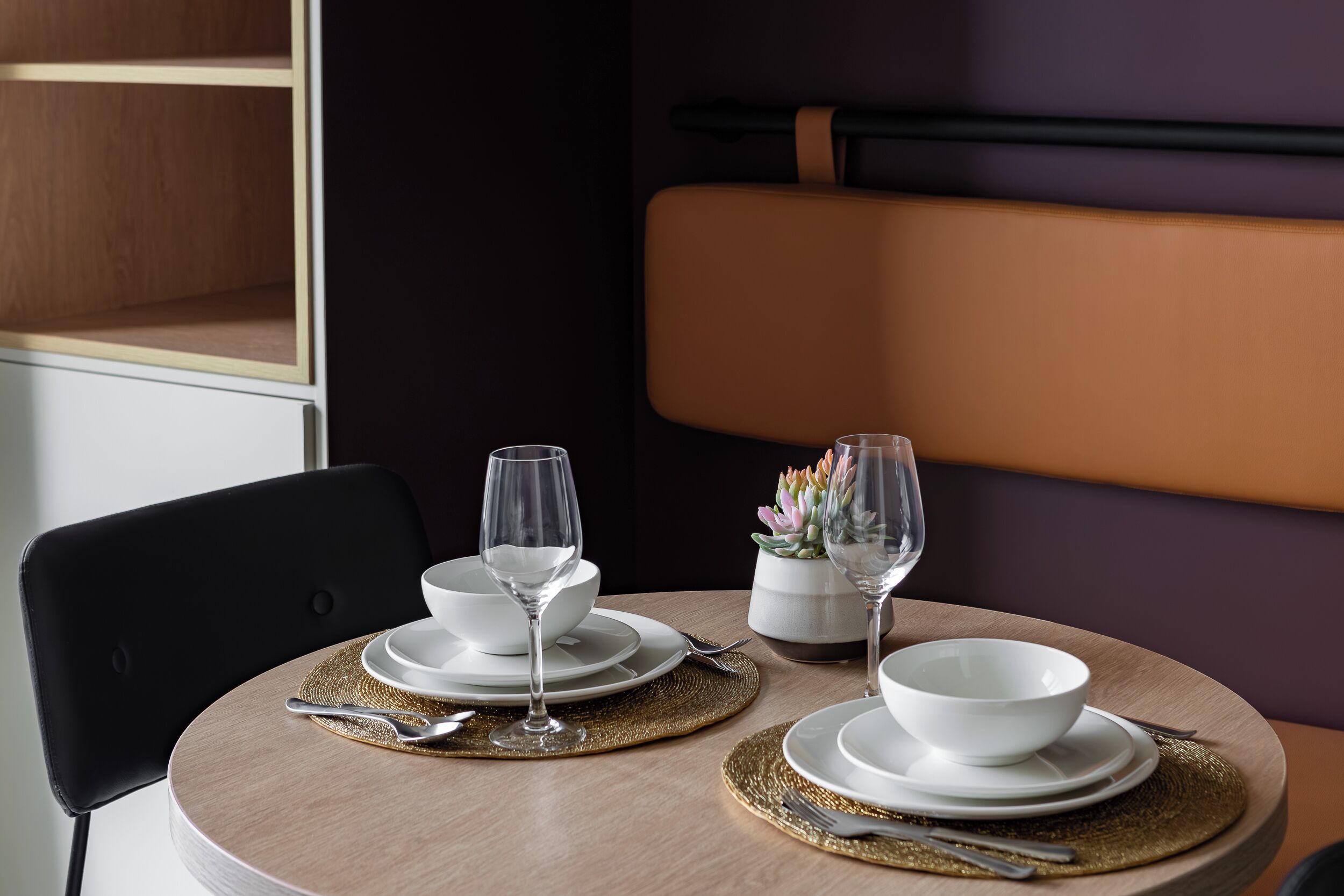 A dining table set with white bowls, glasses, and cutlery on gold placemats, with a chair and a potted plant in the background.