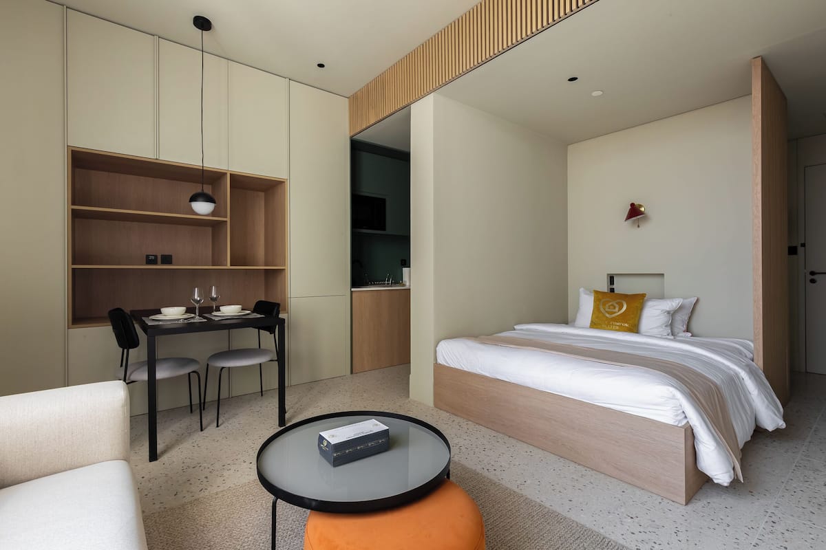 A modern hotel room with a bed, a desk with chairs, a small table, and built-in shelving.