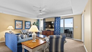 Un salon comprenant un canapé, un fauteuil, une table, une lampe, un téléviseur et un balcon avec des meubles d’extérieur.