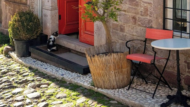 A stone building with a red door, a small outdoor seating area with a table and chairs, and a sign above the door. - Yund 1922 (Ayvalik)