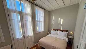 A bedroom with a bed, a nightstand, a lamp, and a window with curtains. - Yund 1922 (Ayvalik)