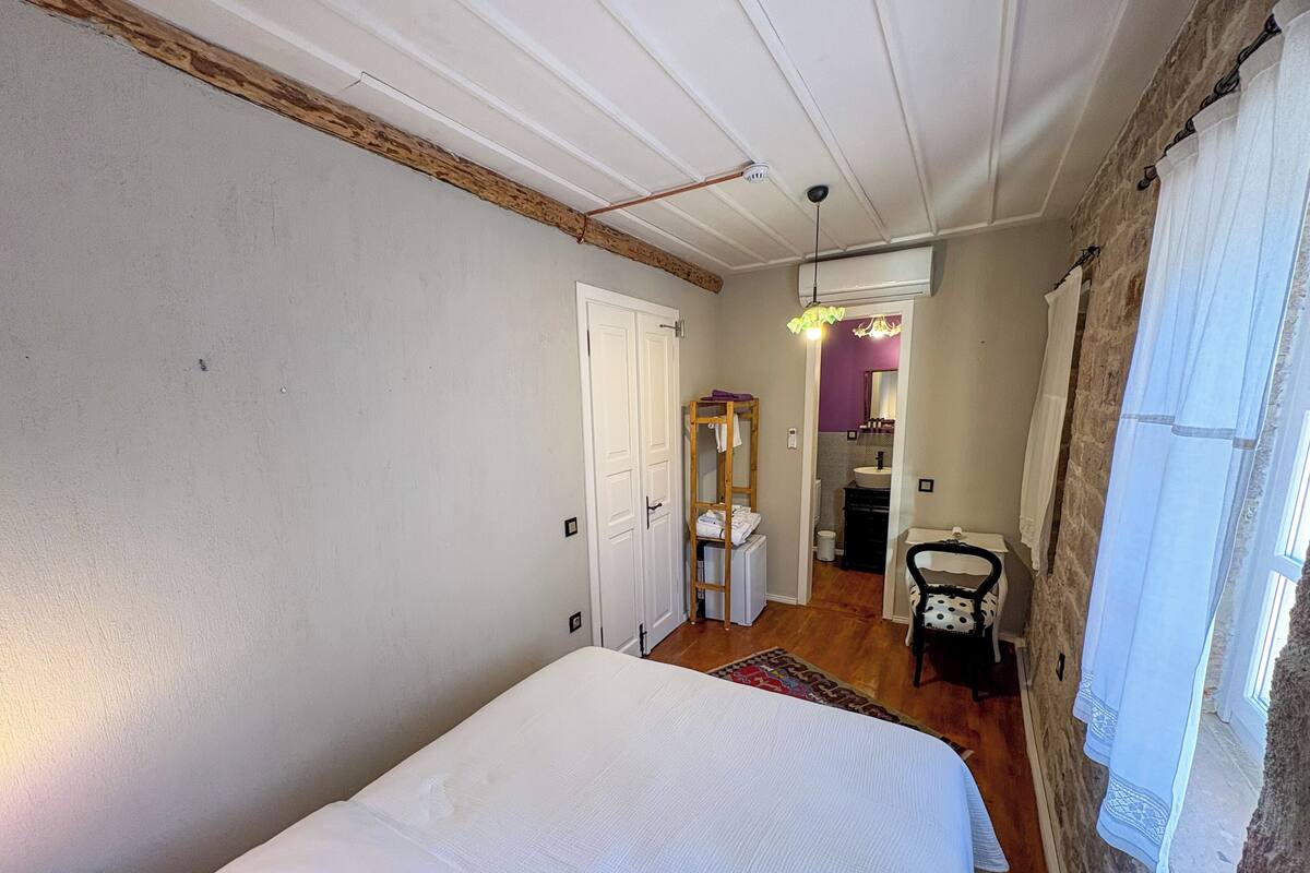 Un dormitorio con cama, techo de vigas de madera, baño con lavabo y espejo, y una pequeña mesa con silla.