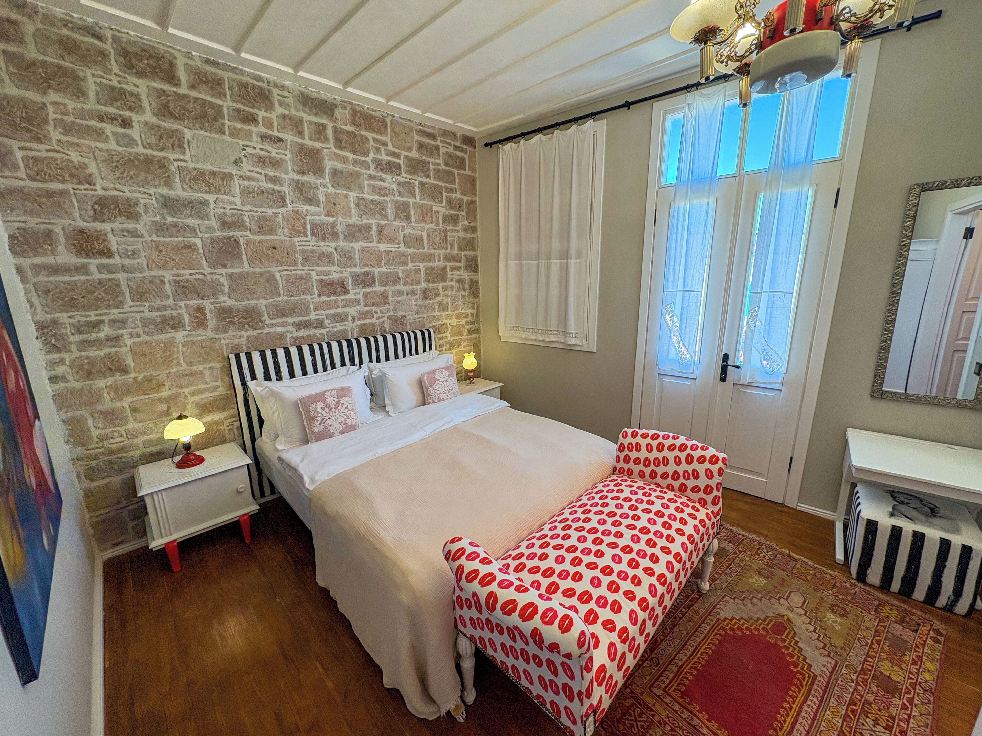 A bedroom with a stone wall, a bed with a striped headboard, a bedside table with a lamp, a red and white patterned bench, and a large mirror.