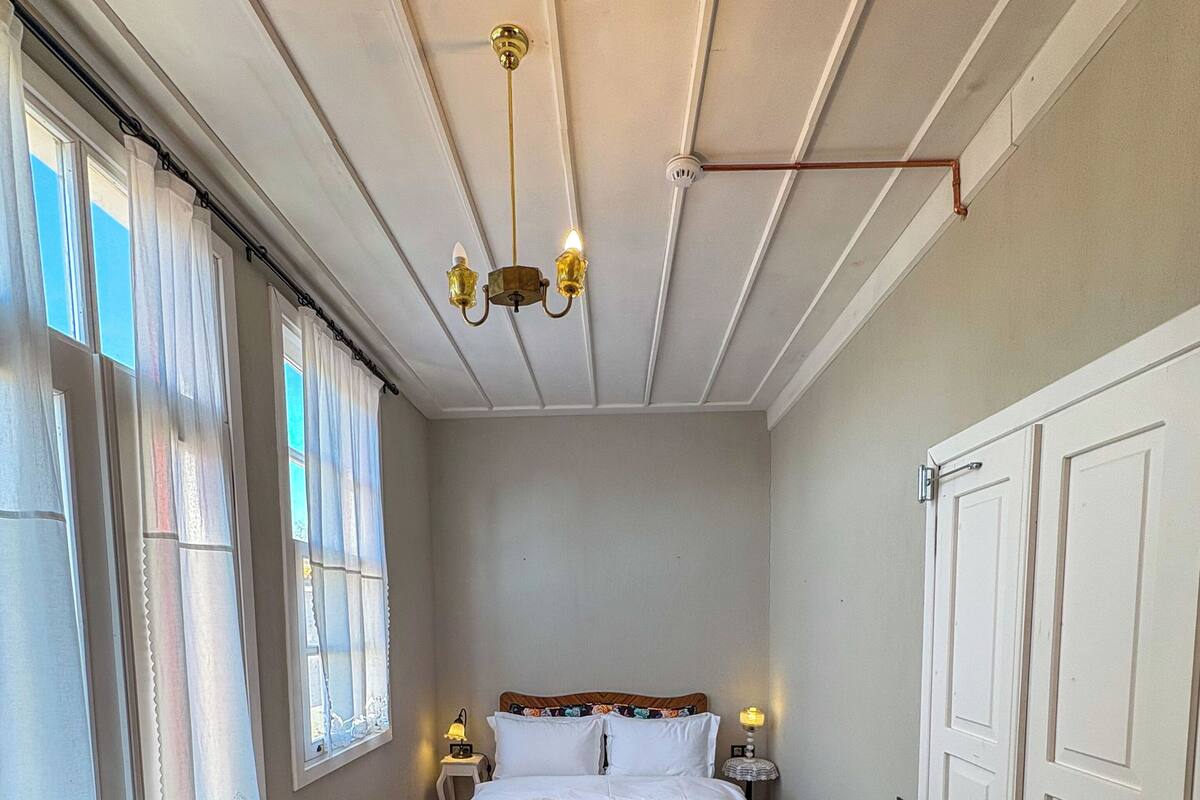 Un dormitorio con cama, dos mesitas de noche con lámparas, una araña, una puerta y una ventana con cortinas.