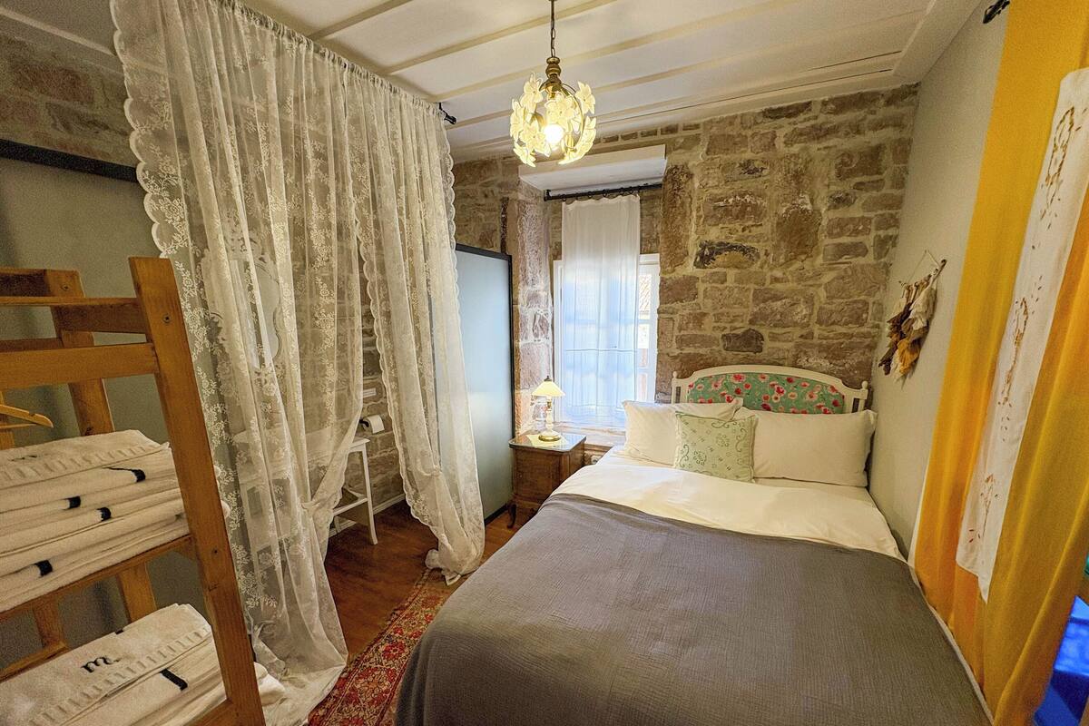 Un dormitorio con pared de piedra, cama de madera, mesita de noche, lámpara, lámpara colgante y ventana con cortina.