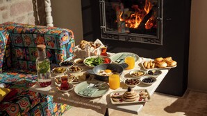 A breakfast table with various dishes and a bottle, set against a backdrop of a fireplace and a colorful sofa. - Yund 1922 (Ayvalik)