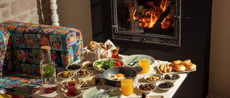 A breakfast table with various dishes and a bottle, set against a backdrop of a fireplace and a colorful sofa.