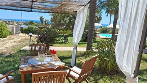 An outdoor seating area with a wooden table, chairs, and a view of the ocean. - La Vela in Sampieri 2 minutes from the sea, villa with pool. (Sicilia)