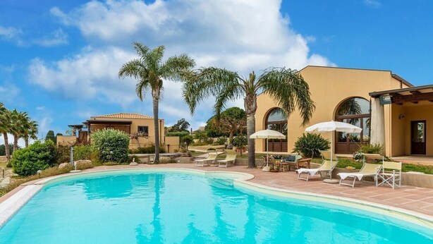 A swimming pool with lounge chairs and umbrellas, surrounded by palm trees and a building with arched windows. - La Vela in Sampieri 2 minutes from the sea, villa with pool. (Sicilia)