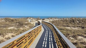 Une promenade en bois menant à la plage, entourée de dunes de sable et d’une végétation clairsemée.