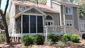 A two-story house with a covered porch, large windows, and a chimney. - Lagoonside Luxury • 5 min Walk to Beach • Pool, Hot Tub & Kiddie Pool (Hilton Head Island)