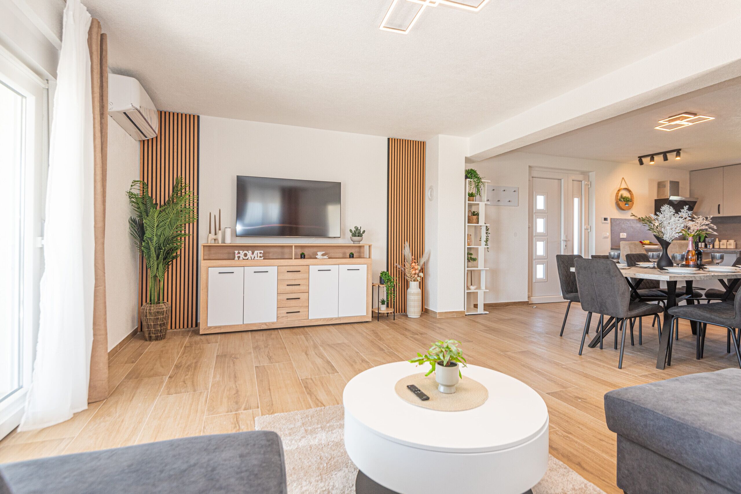 Uma sala de estar moderna com uma televisão de tela plana, um espaço de jantar com mesa e cadeiras, e uma cozinha ao fundo.