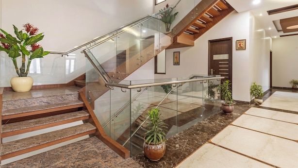 A modern staircase with glass railings and wooden steps, surrounded by potted plants and a polished marble floor. - Miranda by Akama Homes (Noida)
