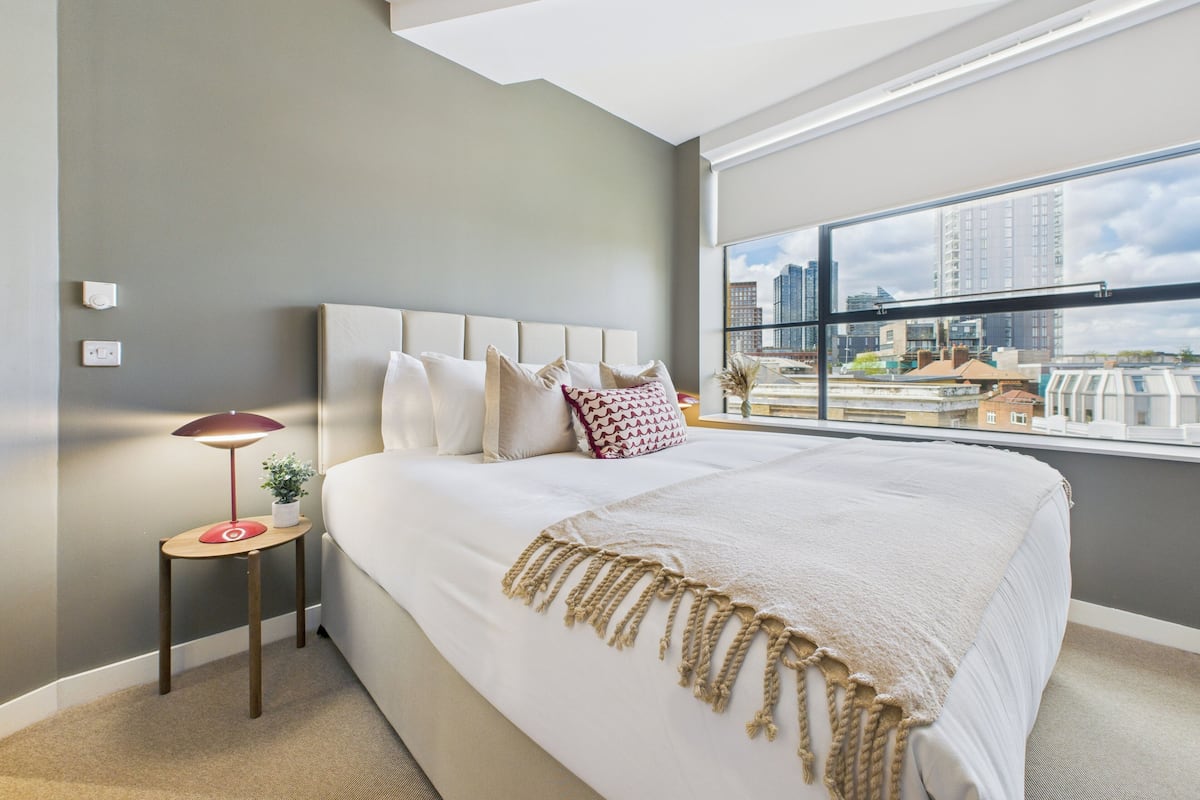 Un dormitorio moderno con una cama grande, una mesita de noche, una lámpara y vistas a la ciudad a través de amplios ventanales.