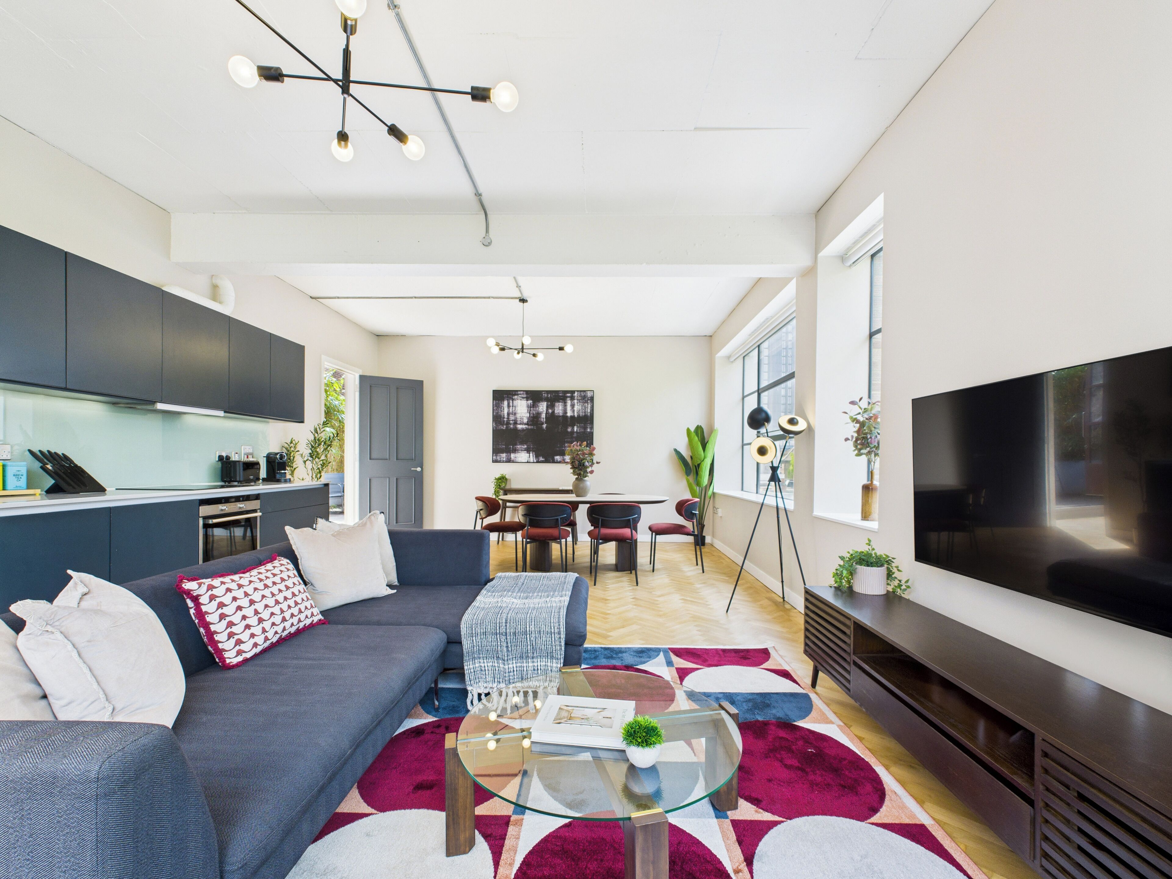 A modern living room with a sofa, coffee table, and TV. There is a kitchen area with cabinets and a dining table.