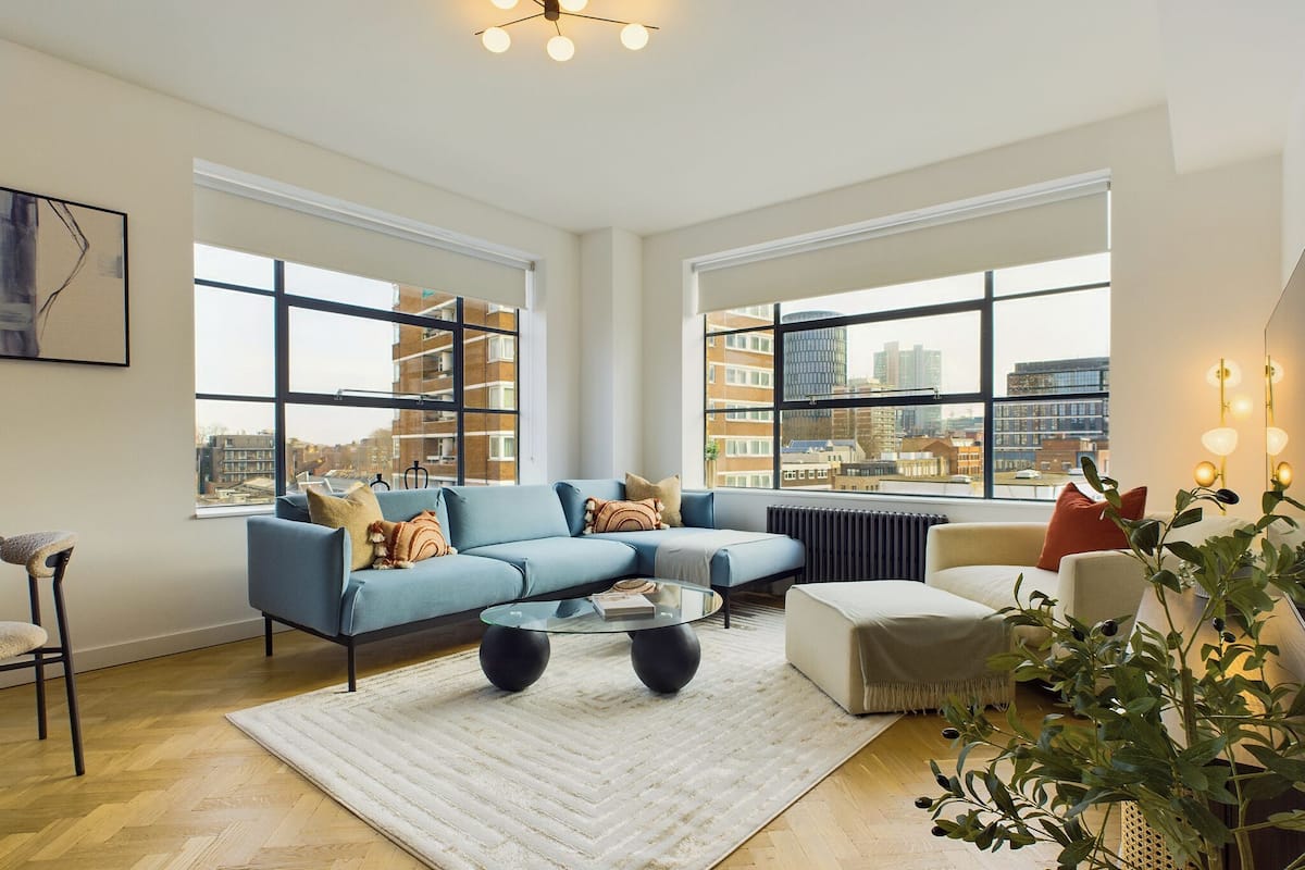 Un salón amplio con un sofá azul, una mesa de centro de vidrio y vistas a la ciudad a través de grandes ventanales.