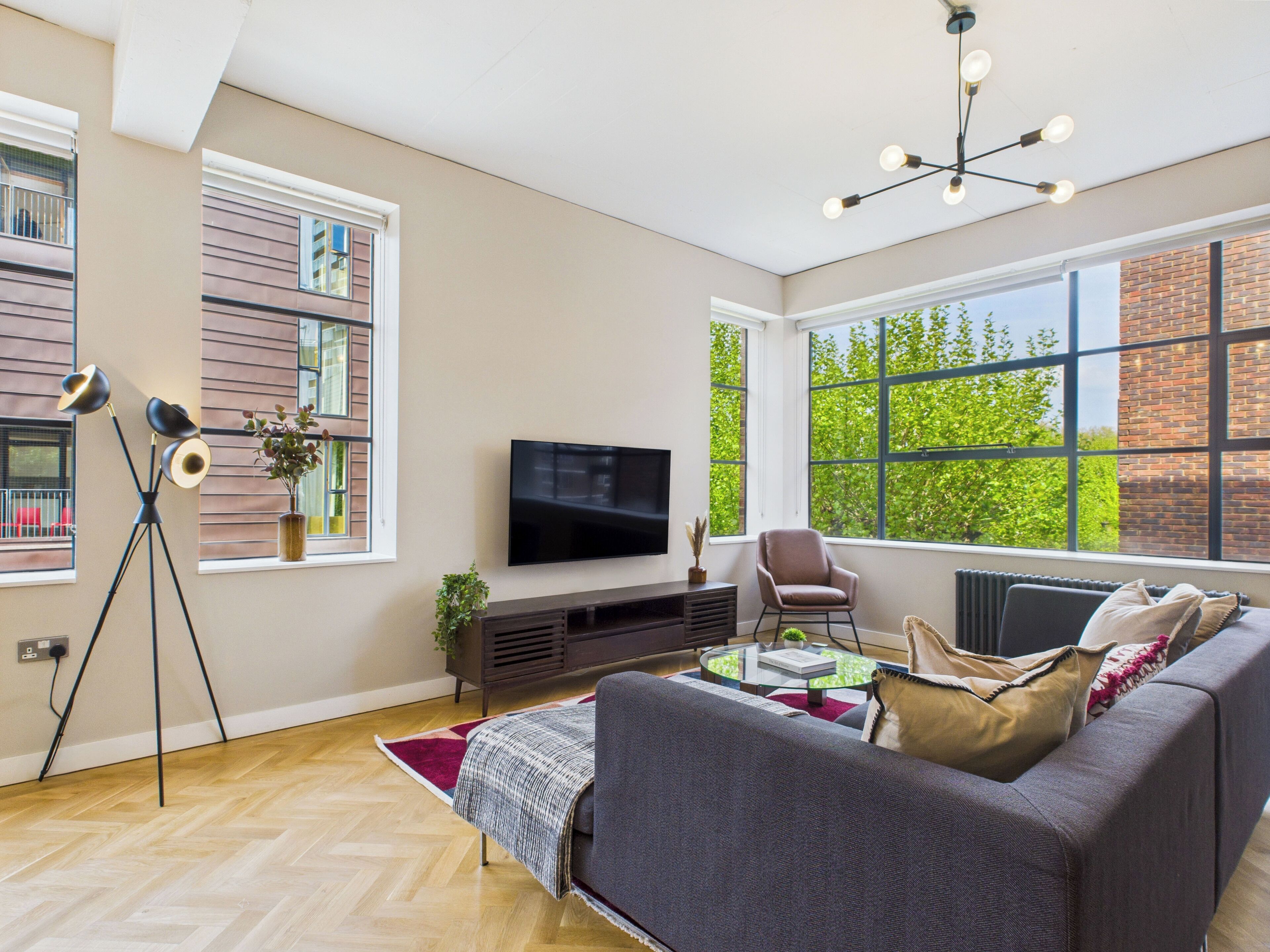 A modern living room with a large TV, comfortable seating, and large windows offering a view of greenery.
