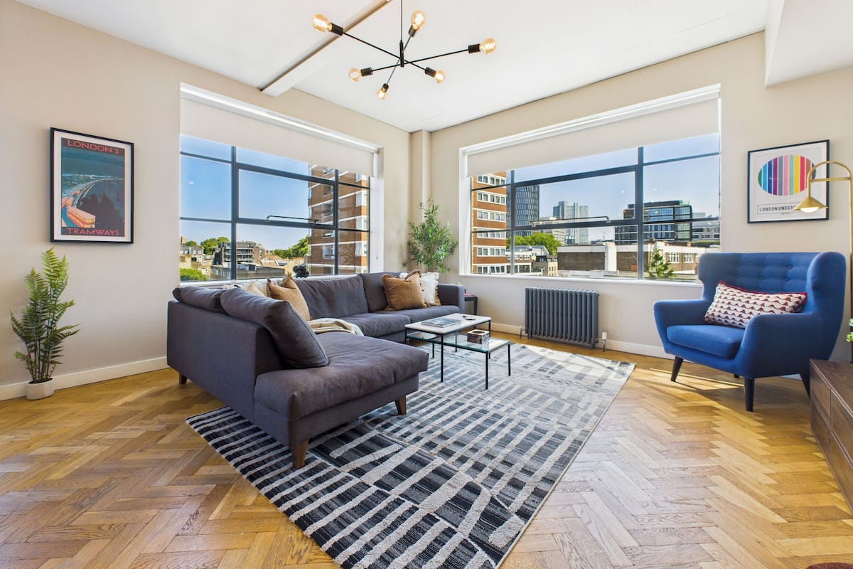 Una sala de estar moderna con sofá, sillón y mesa de centro, ventanas amplias con vistas a la ciudad y un radiador.
