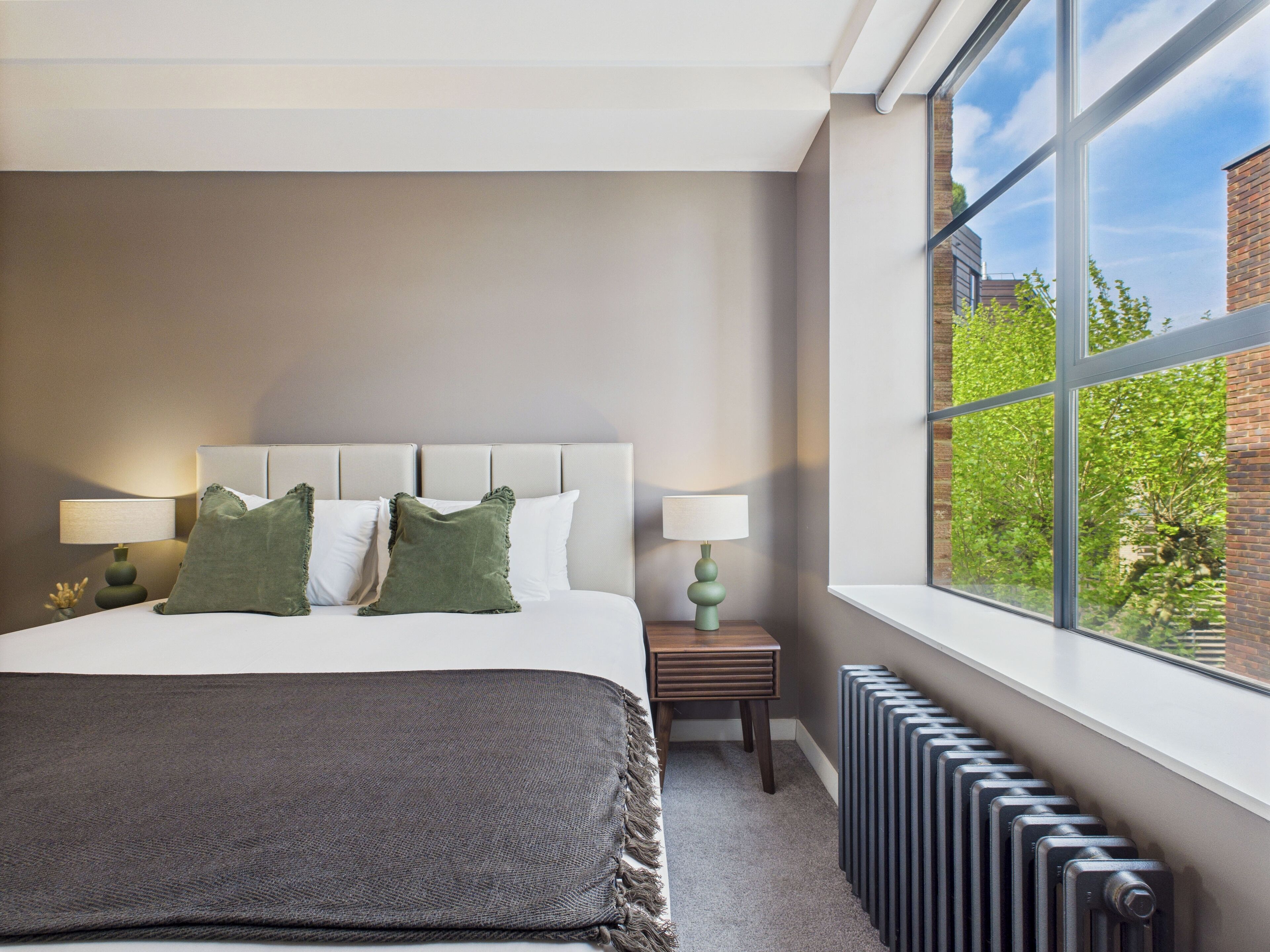Una habitación de hotel moderna con una cama grande, dos lámparas de noche, vistas a zonas verdes y un radiador.