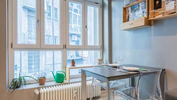 Ein Zimmer mit Tisch, Stühlen, einem Heizkörper, einem Fenster mit Blick auf Gebäude und einem Regal mit Gegenständen.