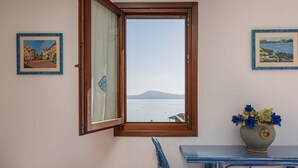 A room with a window overlooking a body of water, a chair, a table, and two framed pictures on the wall. - Loft delle brezze by Wonderful Italy (Olbia)