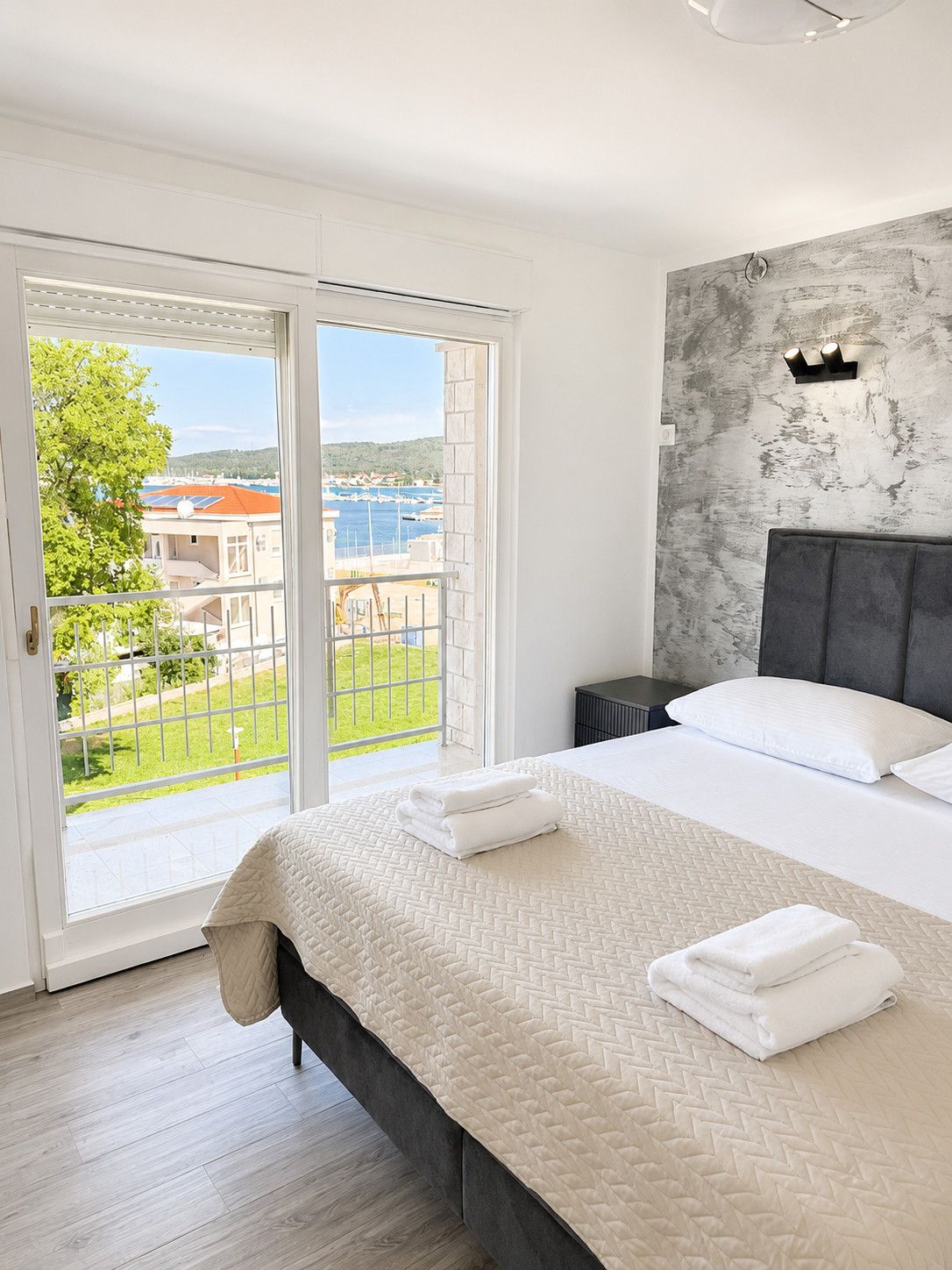 Un dormitorio con cama, mesita de noche, lámpara de pared y vista a un puerto deportivo a través de una puerta corrediza de vidrio.