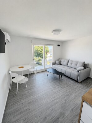 A modern living room with a grey sofa, a round dining table with chairs, and a large window with a view of greenery and buildings. - Mediterraneo apartment A2 (Sukošan)